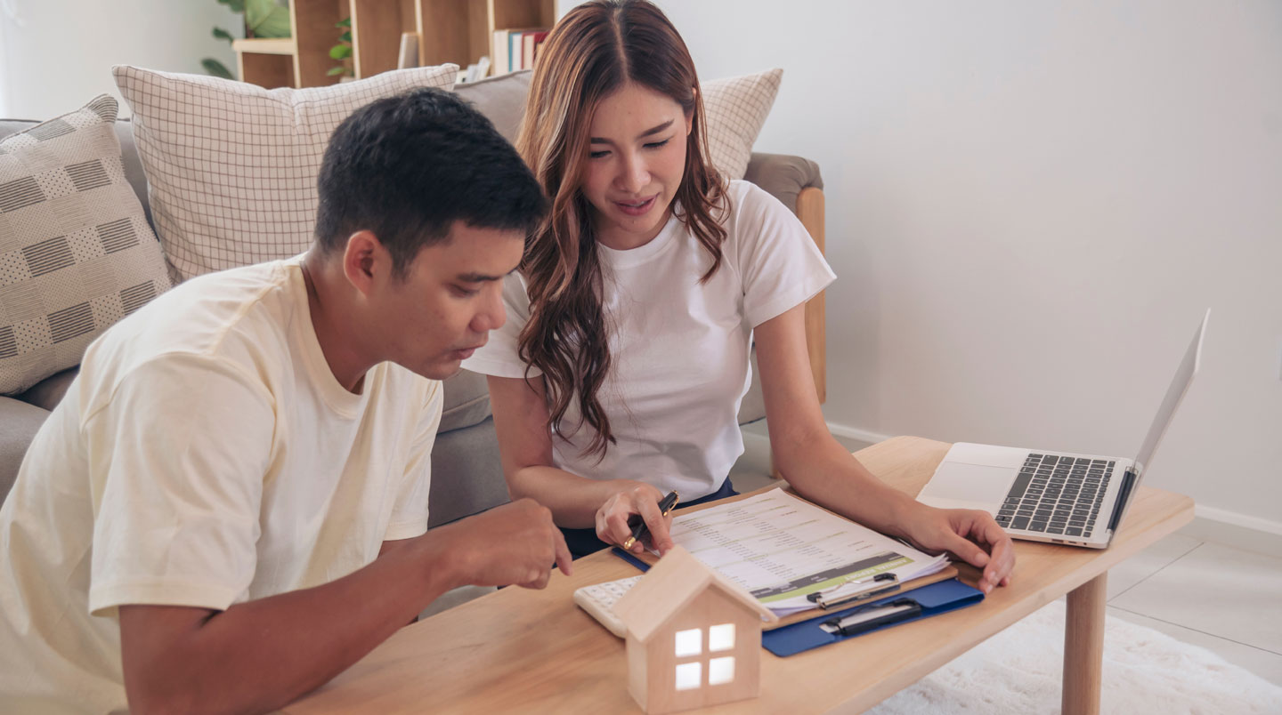 Un couple assis à une table, consultant ensemble des documents d'épargne avec une vue sur leur futur appartement en arrière-plan